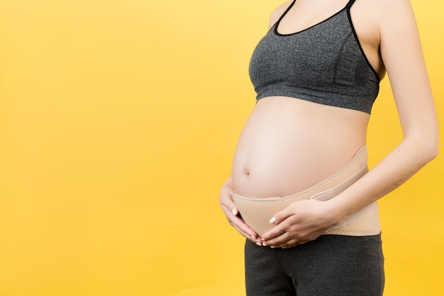Nahaufnahme des Schwangerschaftsverbandes, der auf schwangere Frau gekleidet ist, um Rückenschmerzen an der gelben Oberfläche mit Kopierraum zu reduzieren.
