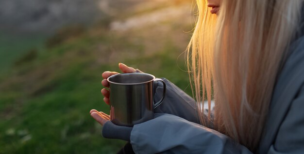 Nahaufnahme des Stahlbechers mit heißem Getränk in den Händen des jungen blonden Mädchens bei susnet.