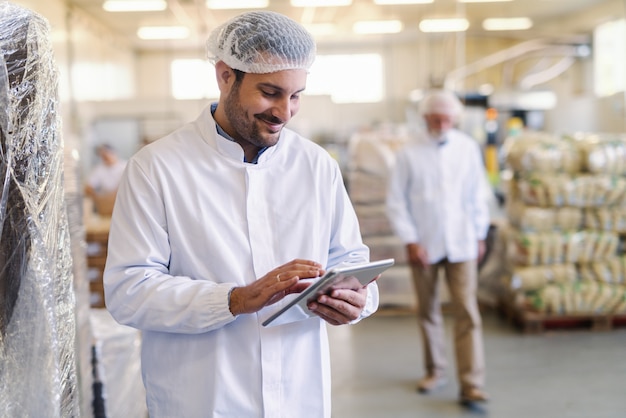 Nahaufnahme des Vorgesetzten in Uniform mit Tablette zur Datenüberprüfung im Stehen in der Lebensmittelfabrik.