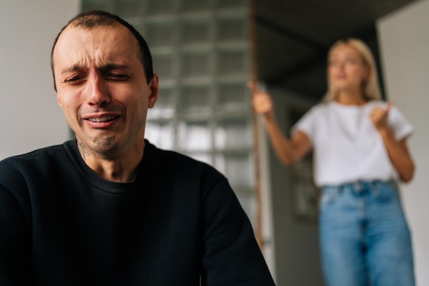 Nahaufnahme des weinenden Mannes, der sich deprimiert und beleidigt fühlt, auf dem Bett sitzt und die wütende, eifersüchtige Frau ignoriert, die argumentiert und dem verärgerten Mann die Schuld für die Probleme zu Hause gibt