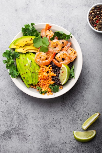 Nahaufnahme, Draufsicht auf Salatschüssel mit Garnelen, Avocado, frischem Grünkohl, Quinoa, roten Linsen, Limette und Olivenöl auf grauem Steinhintergrund. Mittagsschüssel, gesundes, sauberes Essen, Diät oder Ernährungskonzept