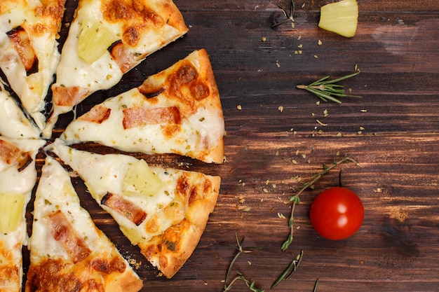 Nahaufnahme Draufsicht Studio schoss acht Scheiben gebackene leckere köstliche italienische hausgemachte Schinkenananas traditionelle hawaiianische Brotkrustenpizza, dekoriert auf rotem Holzkochtisch mit Tomaten und Kräutern