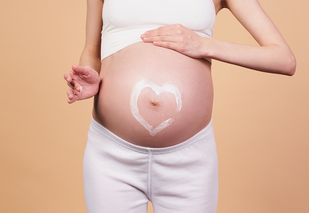 Nahaufnahme, ein schwangeres Mädchen, in einem weißen Sporttop und Leggings, hält ihre Hände auf ihrem nackten Bauch mit herzförmiger Creme darauf. Konzept für Mutterschaft, Geburt, Dehnungsstreifen, schwangere Hautpflege.