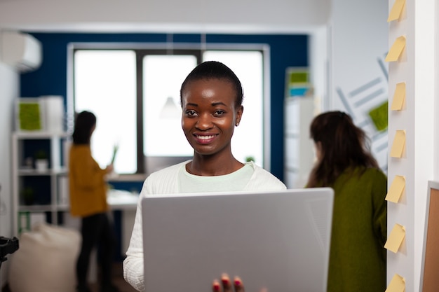 Nahaufnahme einer afrikanischen Frau, die lächelnd in die Kamera schaut, die im Büro der Start-up-Kreativagentur mit Laptop steht und darauf tippt