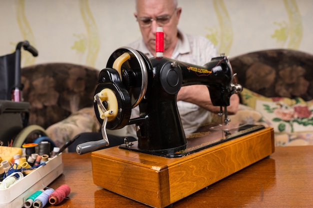 Nahaufnahme einer altmodischen Nähmaschine, umgeben von verschiedenen bunten Fäden - Senior Man hinter Vintage Nähmaschine im Wohnzimmer