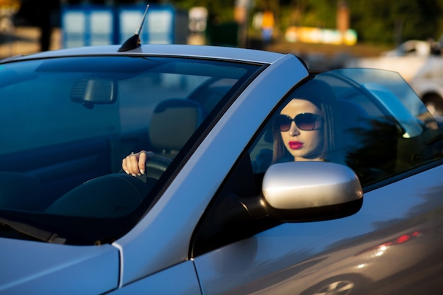 Nahaufnahme einer attraktiven brünetten Frau mit roten Lippen, die eine Sonnenbrille trägt, die ein Cabriolet fährt. Platz für Text