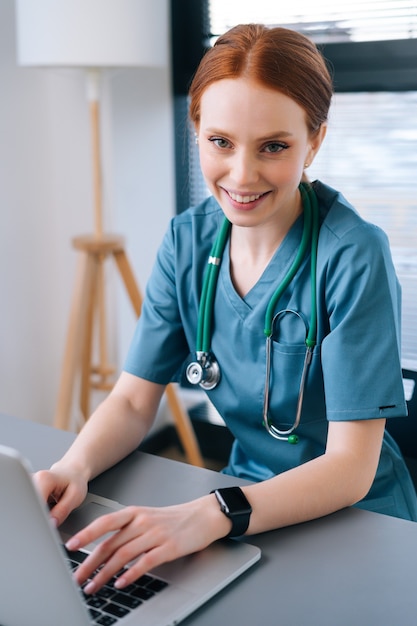 Nahaufnahme einer attraktiven lächelnden jungen Ärztin in blaugrüner medizinischer Uniform, die am Schreibtisch mit Laptop auf dem Hintergrund des Fensters sitzt