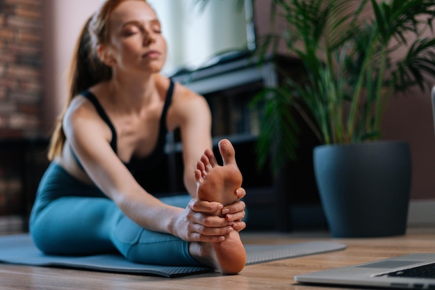 Nahaufnahme einer flexiblen rothaarigen jungen Frau, die beim Stretching auf einer Yogamatte trainiert, während sie online auf dem Laptop Fitnessvideos anschaut Konzept des Sporttrainings einer rothaarigen Dame während der Quarantäne