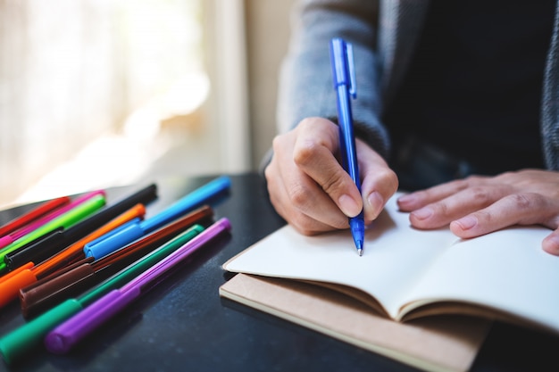 Nahaufnahme einer Frau, die auf ein leeres Notizbuch mit farbigen Stiften auf dem Tisch schreibt