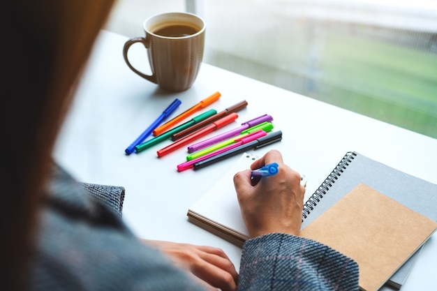 Nahaufnahme einer Frau, die auf ein leeres Notizbuch mit farbigen Stiften und Kaffeetasse auf dem Tisch schreibt