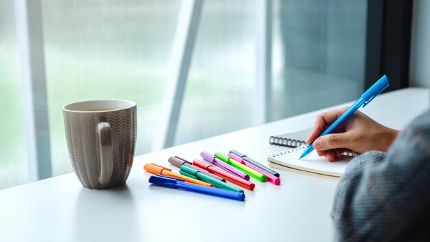 Nahaufnahme einer Frau, die auf ein leeres Notizbuch mit farbigen Stiften und Kaffeetasse auf dem Tisch schreibt