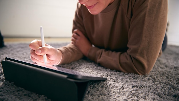 Nahaufnahme einer Frau, die auf einem digitalen Tablet mit einem Stylus-Stift zeichnet, der zu Hause auf dem Teppich liegt