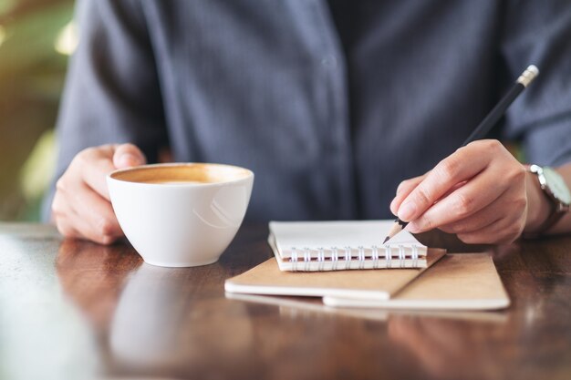 Nahaufnahme einer Frau, die beim Kaffeetrinken auf ein leeres Notizbuch schreibt