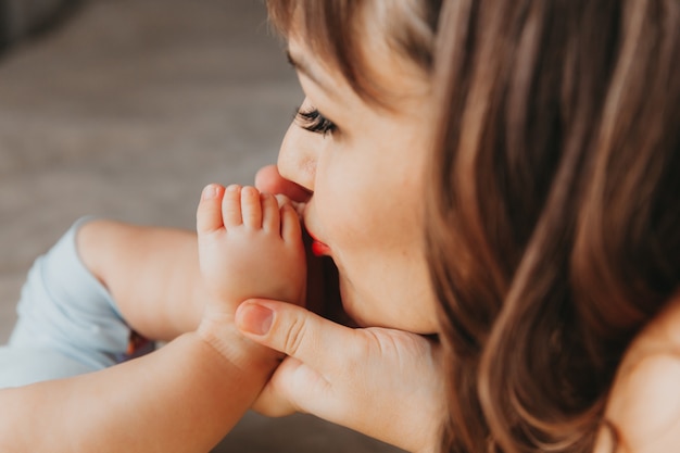 Nahaufnahme einer Frau, die die Beine ihres Babys in der Nähe des Gesichts hält und ihn zärtlich ansieht, während sie Zeit im Schlafzimmer verbringt