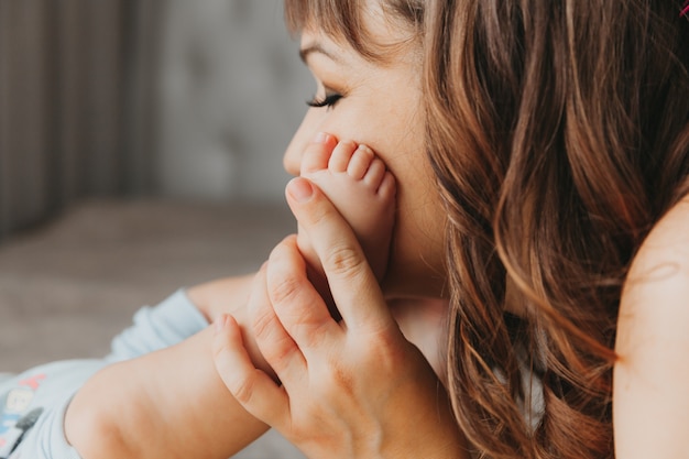 Nahaufnahme einer Frau, die die Beine ihres Babys in der Nähe des Gesichts hält und ihn zärtlich ansieht, während sie Zeit im Schlafzimmer verbringt