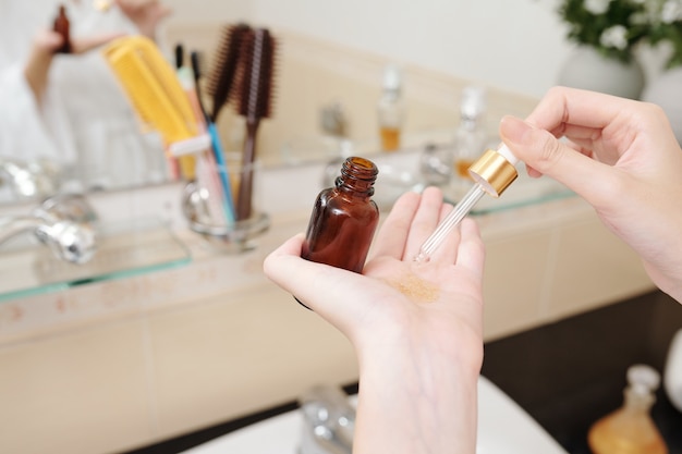 Nahaufnahme einer Frau, die ein paar Tropfen Gesichtsöl in die Hand nimmt, wenn sie am Badezimmerspiegel steht