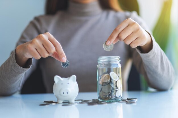Nahaufnahme einer Frau, die eine Münze in ein Sparschwein und ein Glasgefäß steckt, um Geld zu sparen