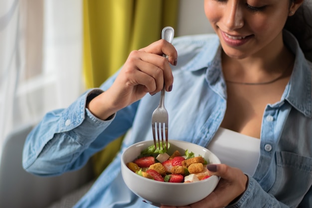 Nahaufnahme einer Frau, die einen Salat isst, den sie auf einem Sofa in ihrem Zimmer sitzt