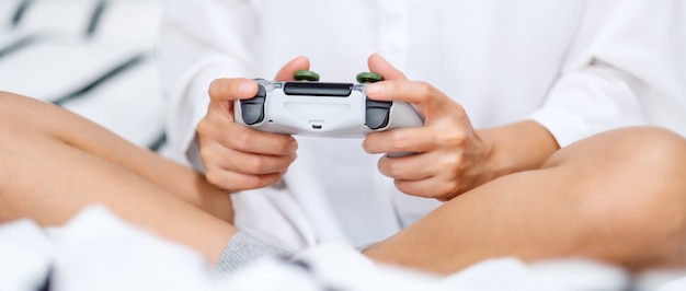 Foto nahaufnahme einer frau, die gamecontroller beim spielen hält und auf einem weißen gemütlichen bett zu hause sitzt