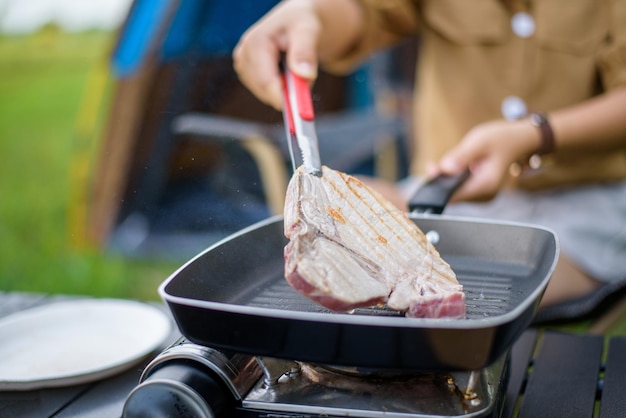 Nahaufnahme einer Frau, die in der Picknickpfanne gegrilltes Schweinefleisch grillt und Essen kocht, während sie auf einem Stuhl auf dem Campingplatz sitzt
