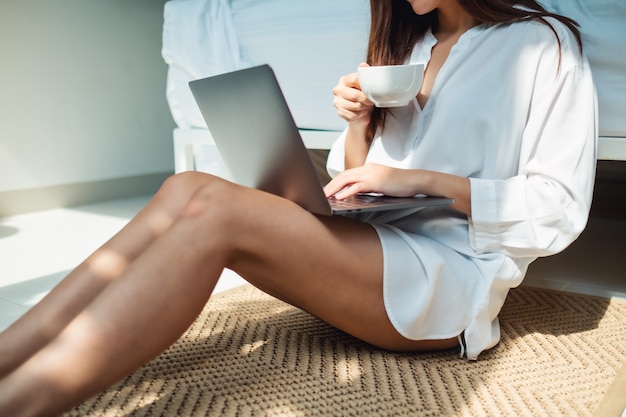 Nahaufnahme einer frau, die laptop-computer benutzt und arbeitet, während kaffee trinkt und im schlafzimmer zu hause sitzt