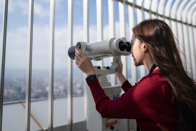 Nahaufnahme einer Frau, die mit dem Teleskop (dem Fernglas) auf der Oberseite des Gebäudes auf die Stadt schaut.