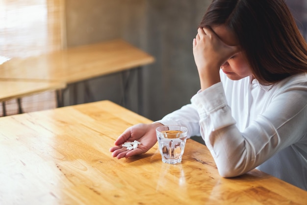 Nahaufnahme einer Frau mit Kopfschmerzen, die eine weiße Pille mit einem Glas Wasser auf dem Tisch hält