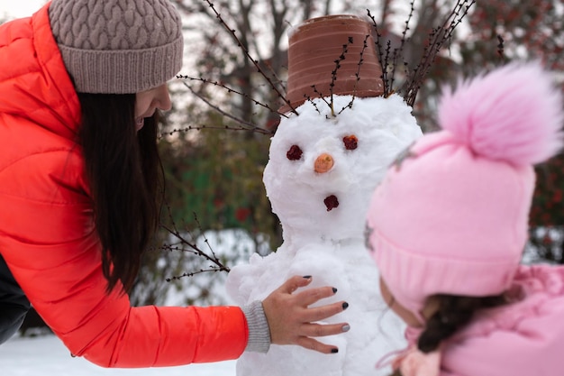 Nahaufnahme einer Frau und eines kleinen Mädchens, die draußen tagsüber im Winter einen Schneemann mit Schnee bauen, während sie ...