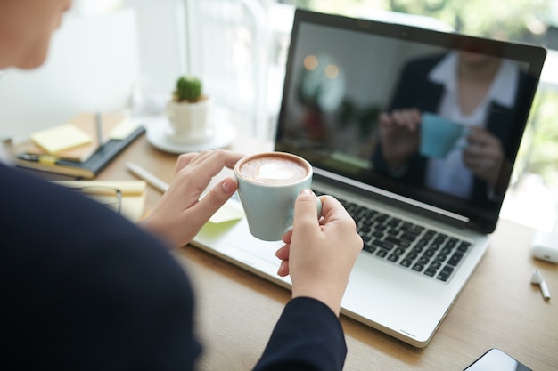 Nahaufnahme einer Geschäftsfrau, die eine Tasse köstlichen Cappuccino trinkt, wenn sie eine kurze Pause macht, nachdem sie E-Mails an Geschäftspartner gesendet hat