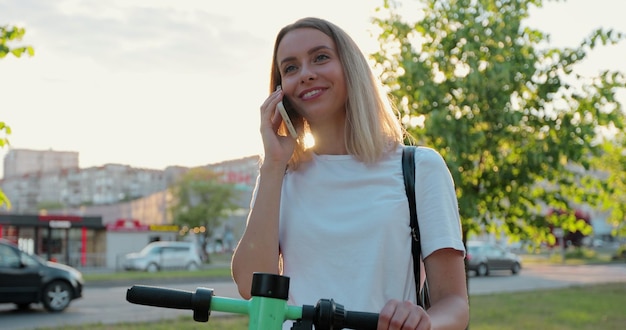 Nahaufnahme einer glücklichen Frau steht mit einem modernen Elektroroller, der bei Sonnenuntergang in der Stadtstraße telefoniert Hübsches Mädchen, das im Sommer auf der Straße einen Elektroroller fährt und telefoniert