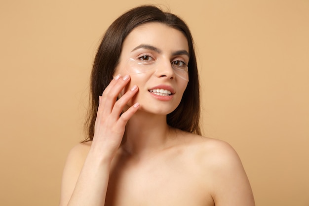 Nahaufnahme einer halbnackten Frau mit perfekter Haut, nackte Make-up-Patches unter den Augen isoliert auf beige pastellfarbener Wand