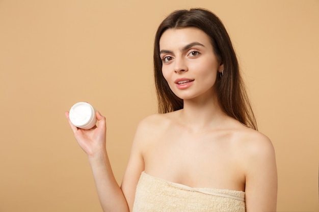 Nahaufnahme einer halbnackten Frau mit perfekter Haut, nacktem Make-up, das Gesichtscreme auf beige pastellfarbener Wand isoliert