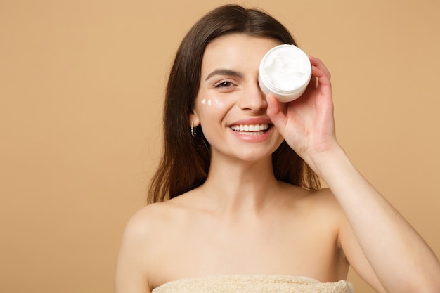 Nahaufnahme einer halbnackten Frau mit perfekter Haut, nacktem Make-up, das Gesichtscreme auf beige pastellfarbener Wand isoliert