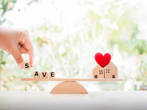 Nahaufnahme einer Hand, die einen Holzblock mit dem Wort „SAVE“ und einem Miniaturhaus auf der Waage hält. Das Konzept der Steuerzahlung für Haus und Eigentum