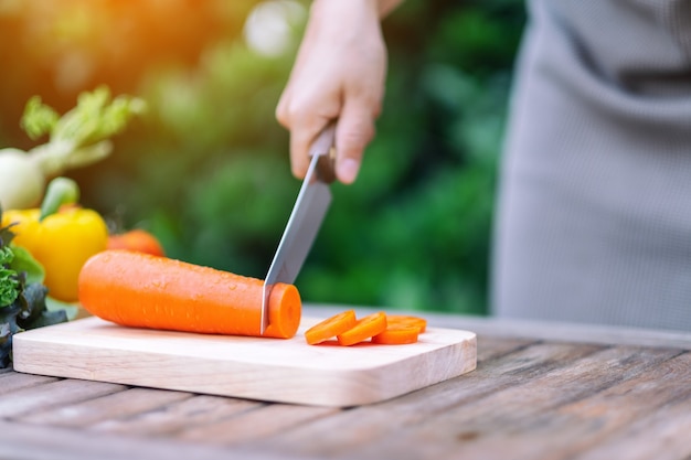 Nahaufnahme einer Hand, die Karotten mit dem Messer auf Holzbrett schneidet und hackt