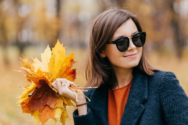 Nahaufnahme einer hübschen Frau mit Haaren, die eine Sonnenbrille trägt, hat einen Spaziergang an einem sonnigen Tag im Herbst im Park und trägt gelbe Blätter, die nachdenklich beiseite schauen Menschen Saison und Lifestyle-Konzept