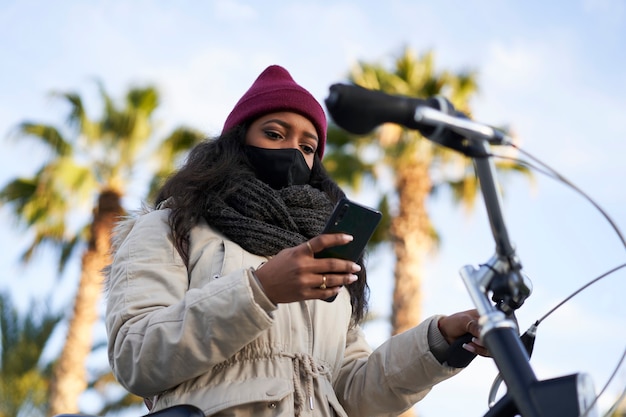 Nahaufnahme einer jungen afroamerikanischen Frau auf ihrem Fahrrad, Winterkleidung tragend, mit ihrem Smartphone.