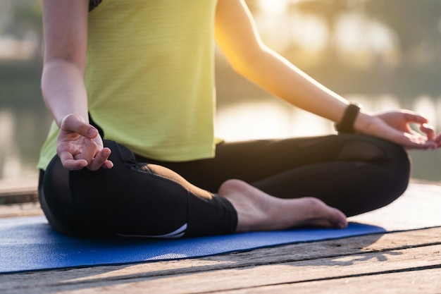 Nahaufnahme einer jungen asiatischen schönen Frau, die Yoga praktiziert und morgens im Lotussitz im Freien neben dem See meditiert, um sich zu entspannen und zu beruhigen. Harmonie- und Meditationskonzept.