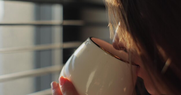 Foto nahaufnahme einer jungen frau, die bei sonnenaufgang in der nähe eines fensters mit jalousien heißen kaffee oder tee trinkt