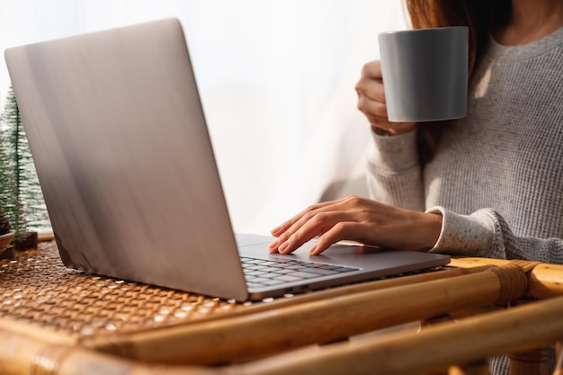 Nahaufnahme einer jungen Frau, die einen Laptop verwendet und daran arbeitet, während sie zu Hause Kaffee trinkt