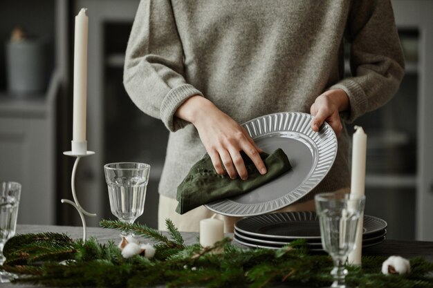 Nahaufnahme einer jungen Frau, die Teller auf dem weihnachtlich dekorierten Esstisch mit Tannenzweigen aufstellt ...