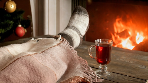 Nahaufnahme einer jungen Frau in Wollsocken, die unter einer Decke neben einem brennenden Kamin mit einem Glas Glühwein liegt?