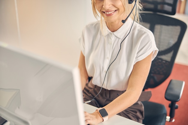 Nahaufnahme einer jungen Frau mit Headset, die mit Desktop-PC am Tisch sitzt und lächelt