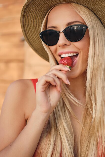 Nahaufnahme einer jungen, fröhlichen, blonden Frau mit Sonnenbrille und Hut, die glücklich Erdbeeren am Strand isst