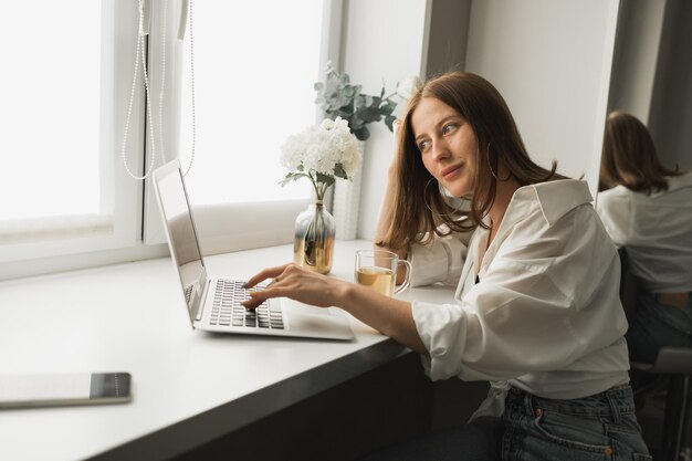 Nahaufnahme einer jungen hübschen Frau, die während der Pause Tee trinkt, nachdem sie online auf Laptop w getippt und gesurft hat