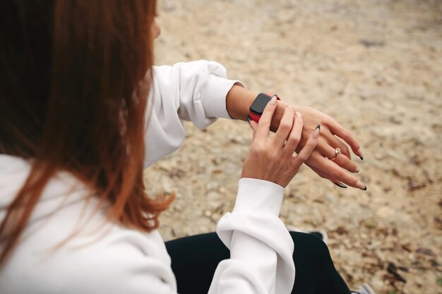 Nahaufnahme einer jungen rothaarigen Frau, die nach dem Training oder dem Laufen im Freien auf dem Strand sitzt und eine weiße Kapuzen-Check-Smartwatch trägt