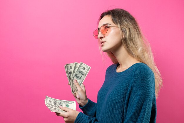 Nahaufnahme einer jungen schönen Frau in einem blauen Pullover in Gläsern mit US-Dollar-Geld in der Hand