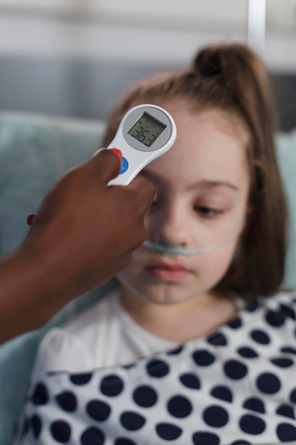 Nahaufnahme einer Kinderkrankenschwester, die die Körpertemperatur eines kranken kleinen Mädchens mit einem Thermometer misst. Das medizinische Personal der Kindergesundheitsklinik untersucht die Krankheitsentwicklung eines kranken Kindes, das in Behandlung ist