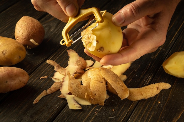 Nahaufnahme einer Kochhand, die mit einem Kartoffelschälter rohe Kartoffeln schält, um das Mittagessen vorzubereiten Gemüse-Menü-Konzept für Restaurant oder Hotel auf schwarzem Hintergrund