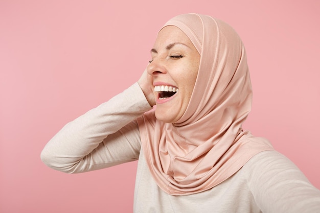 Nahaufnahme einer lachenden lustigen jungen arabischen muslimischen Frau in hijab leichter Kleidung, die isoliert auf rosa Hintergrund posiert. Menschen religiöses Lebensstilkonzept. Mock-up-Kopienbereich. Selfie-Aufnahme auf dem Handy machen.